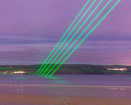 Five rays of green laser lights brake over the surface of the water in a pink coloured sunset over the sea